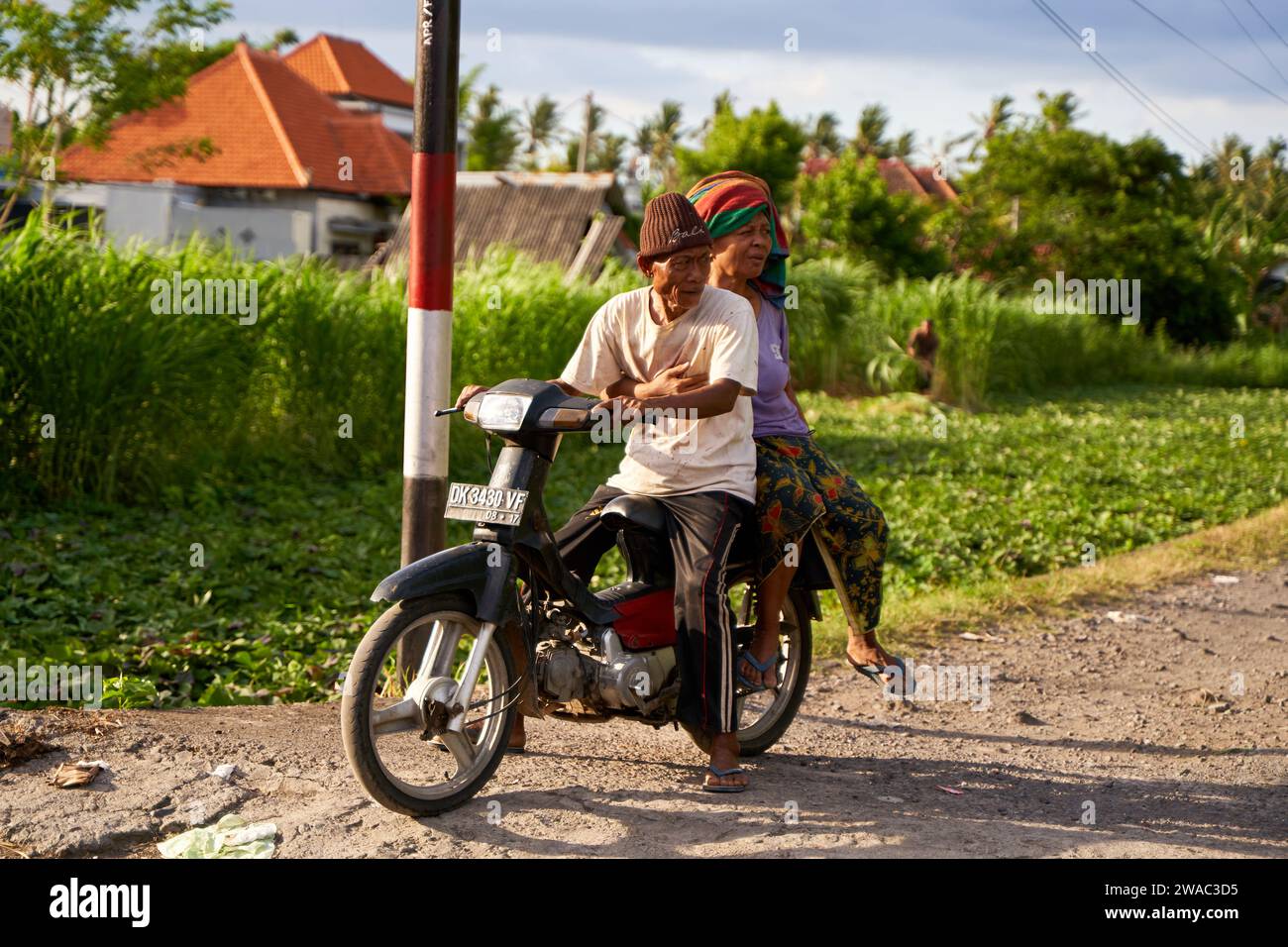 A picturesque local elderly couple riding a motorcycle on a road in a village in Bali. Bali, Indonesia - 12.10.2022 Stock Photo