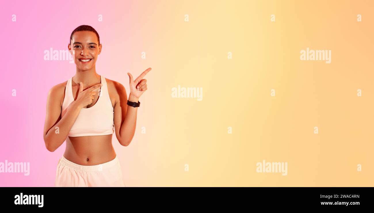 An engaging woman with a shaved head points to her left with a bright smile, dressed in a white sports bra Stock Photo