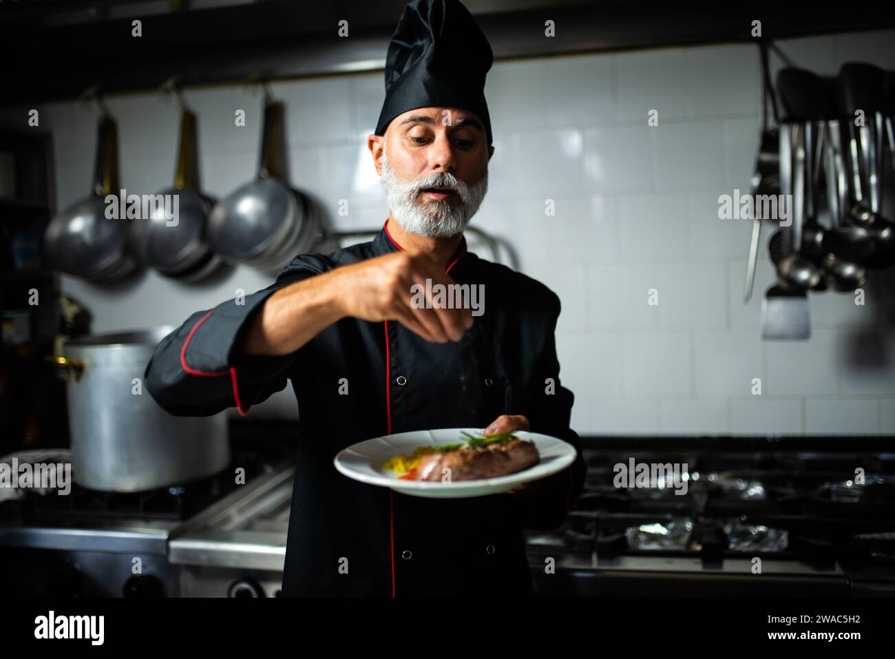 Mature chef in uniform focuses on garnishing a plated meal in a commercial kitchen setting Stock Photo