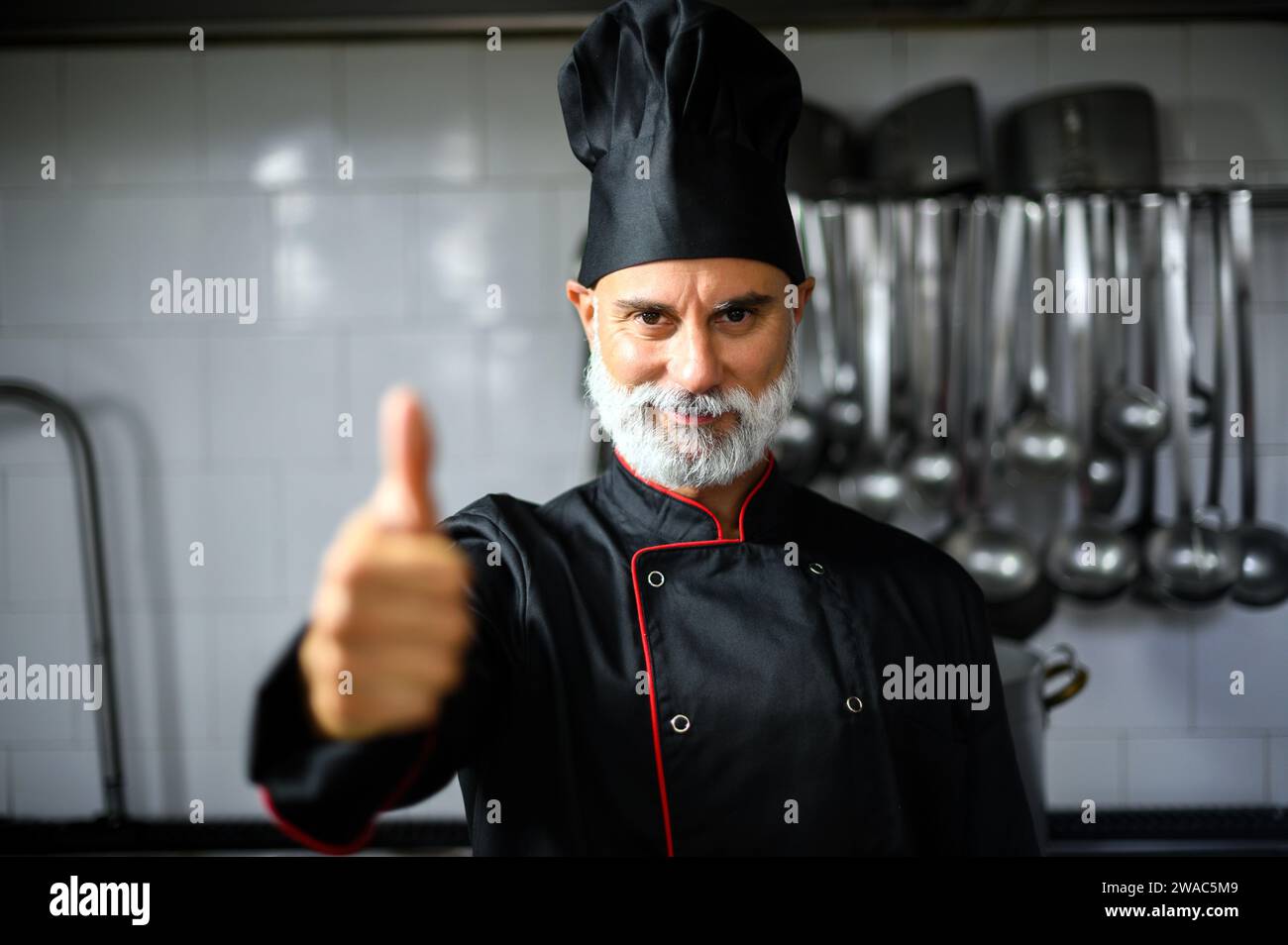 Mature chef in black coat giving thumbs up in his kitchen Stock Photo