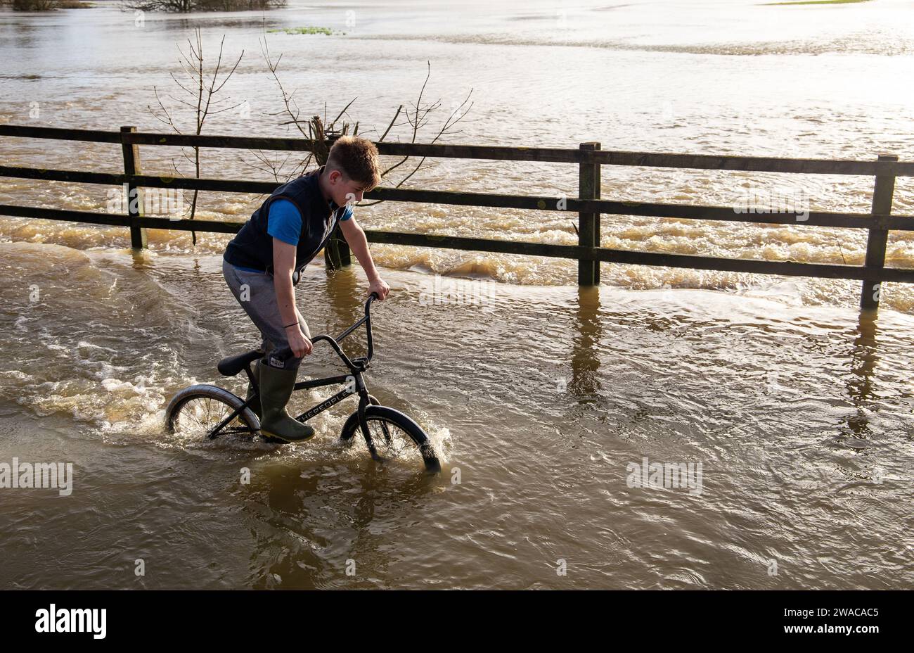 Boy riding bicycle through flood at Lacock after storm Henk 3rd Jan 24 Stock Photo
