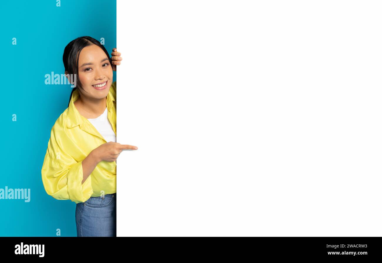 An Asian woman with a bright smile is peeking from behind a blank white vertical banner Stock Photo