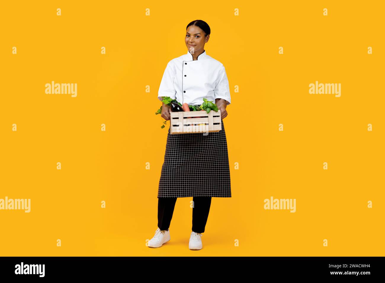 Smiling black female chef presenting wooden crate of organic vegetables ...
