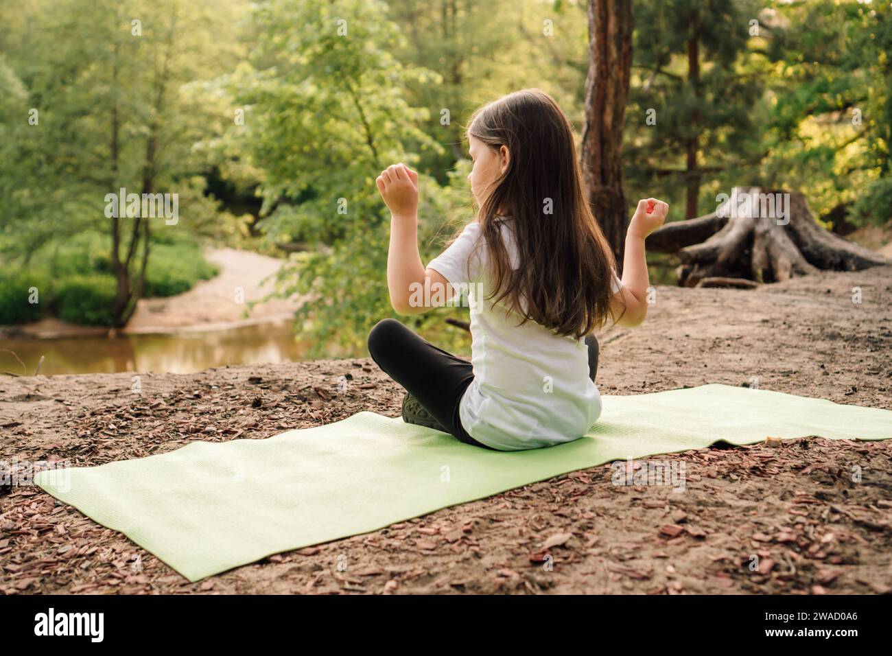 Rear view of female sporty child peacefully sitting on green mat in yoga pose with raised arms. Little girl with long brown hair closed eyes and doing Stock Photo