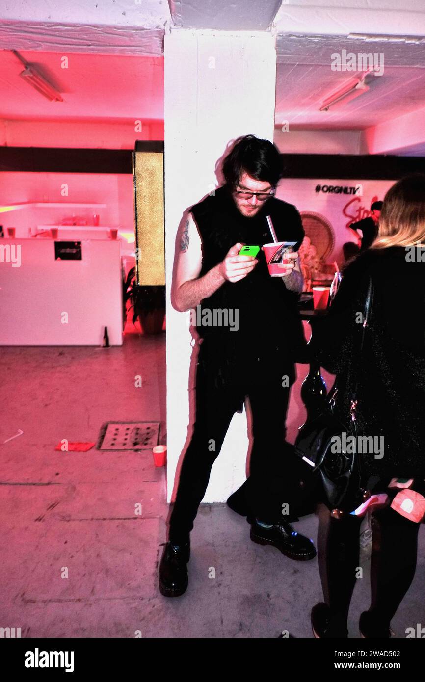 Man dressed in black, black hair, black glasses with a drink in a red paper cup holding a lime green phone at a warehouse party event, pink lighting Stock Photo
