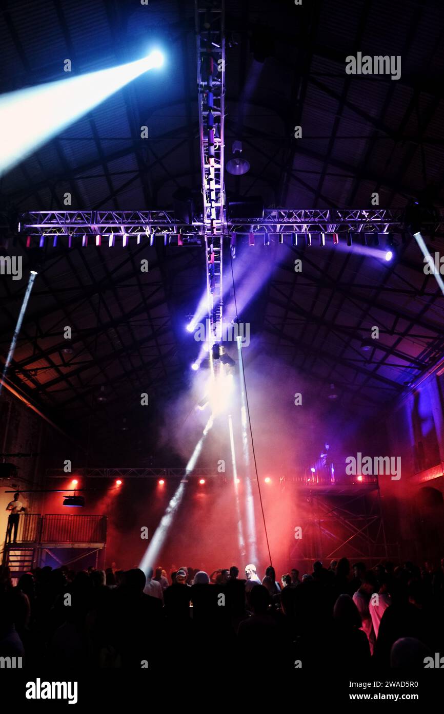 Coloured spotlights, cross boom rigging overhead, a rave party in an industrial warehouse, red purple & blue, smoke haze & tiny people in silhouette Stock Photo