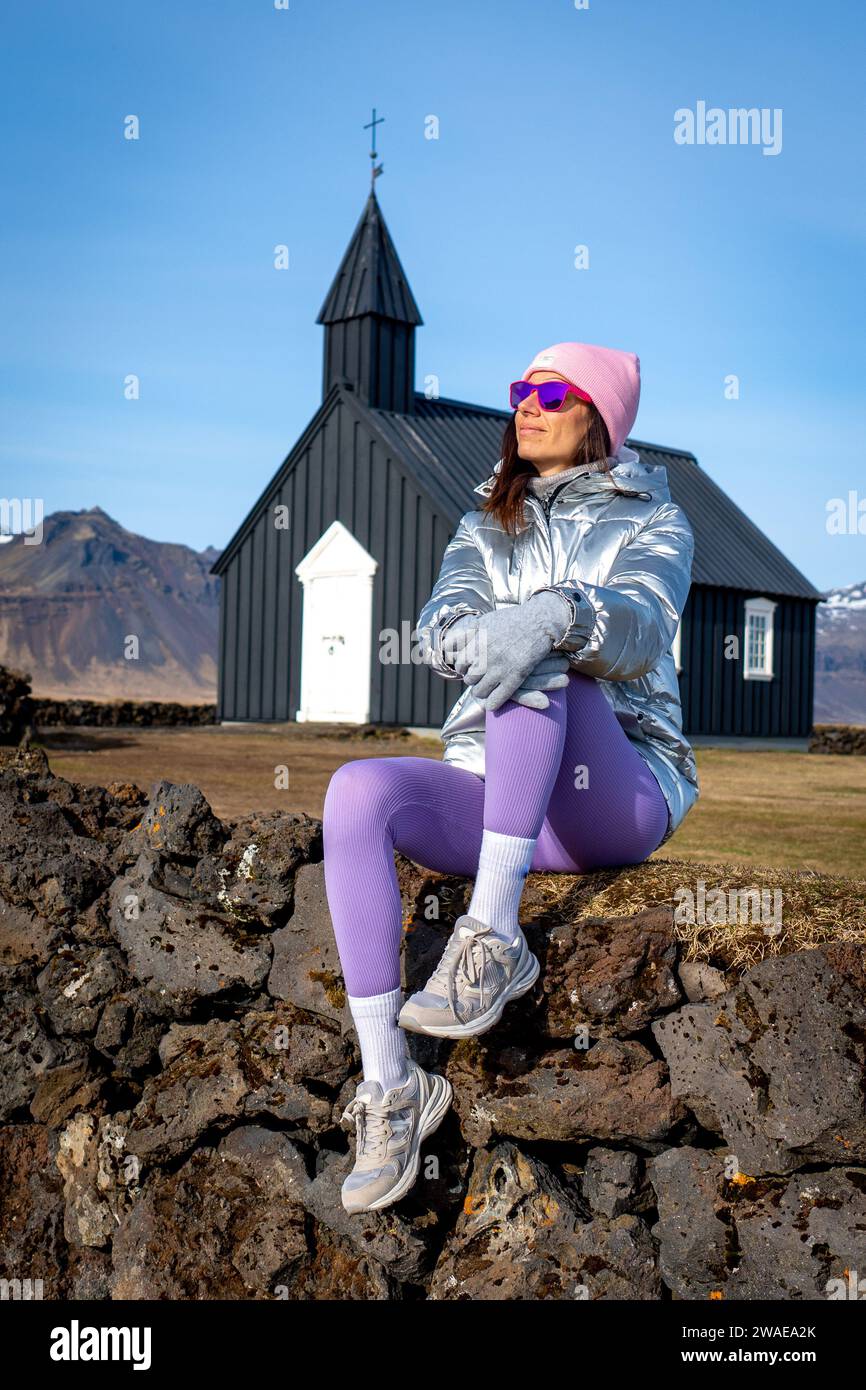 A young woman wearing a purple hat, silver jacket, and blue leggings is seated in a relaxed pose Stock Photo