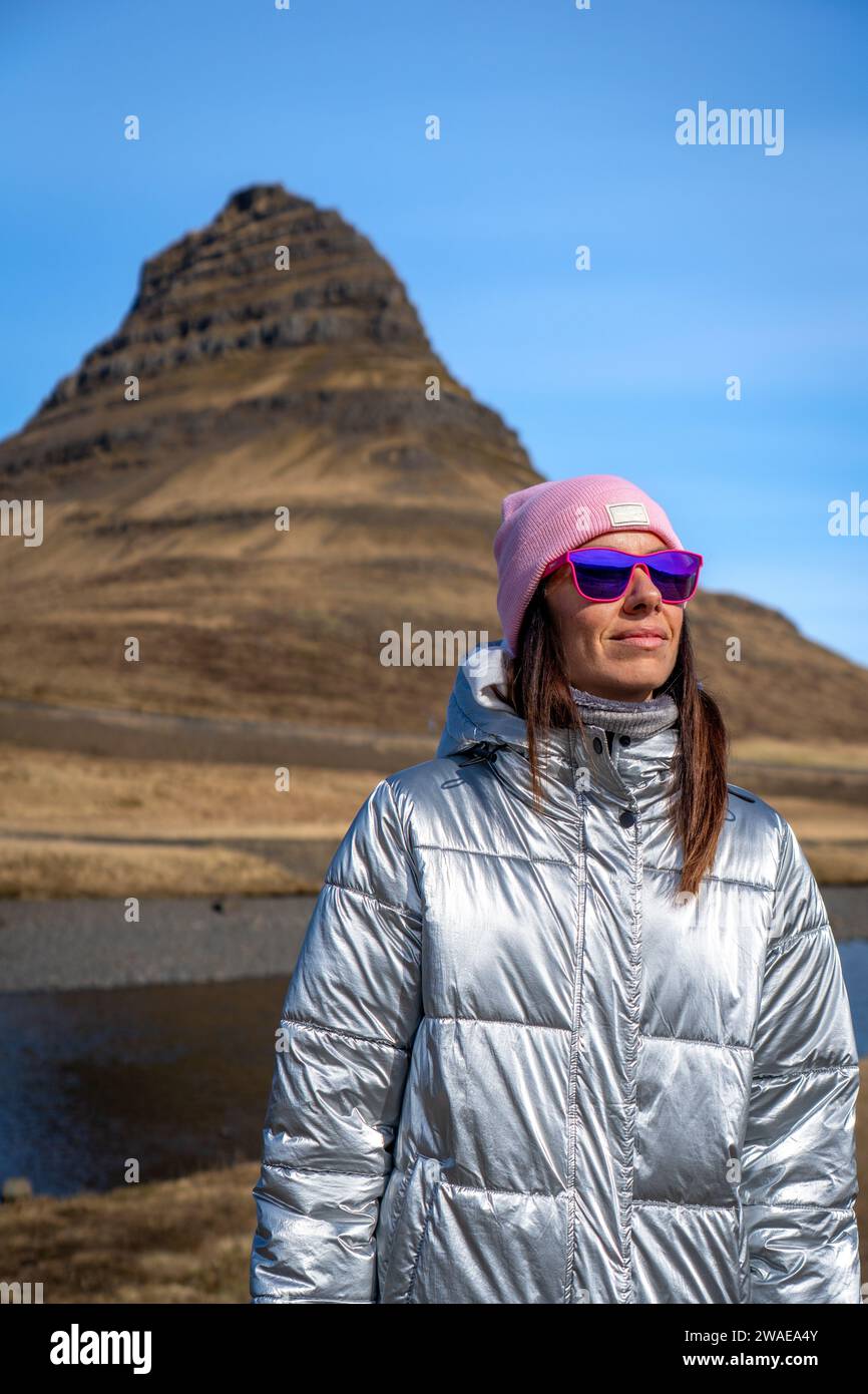 A young female stands in the foreground, wearing a silver puffy jacket and a pink hat, with a stunning alpine mountain range in the background Stock Photo