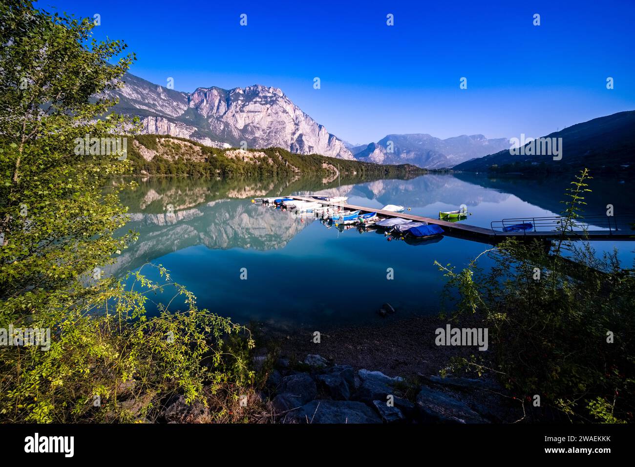 Summit and ridges of the mountain Monte Casale reflecting in the water of the lake Lago di Cavedine, rowing boats attached to a jetty. Stock Photo