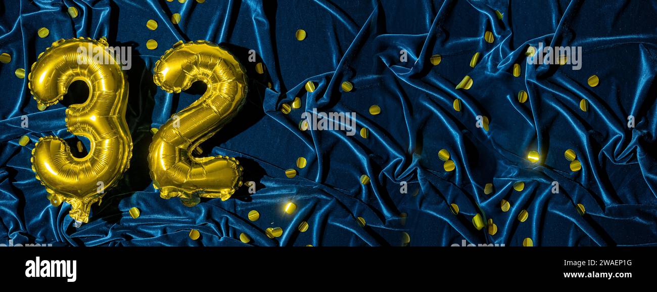 number thirty two, gold foil balloon number on blue velvet background with confetti. Birthday greeting card, inscription 32. Anniversary event. Banner Stock Photo