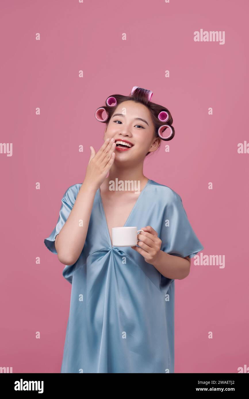 Beautiful young woman in blue dress and hair curlers drinking coffee on pink background Stock Photo