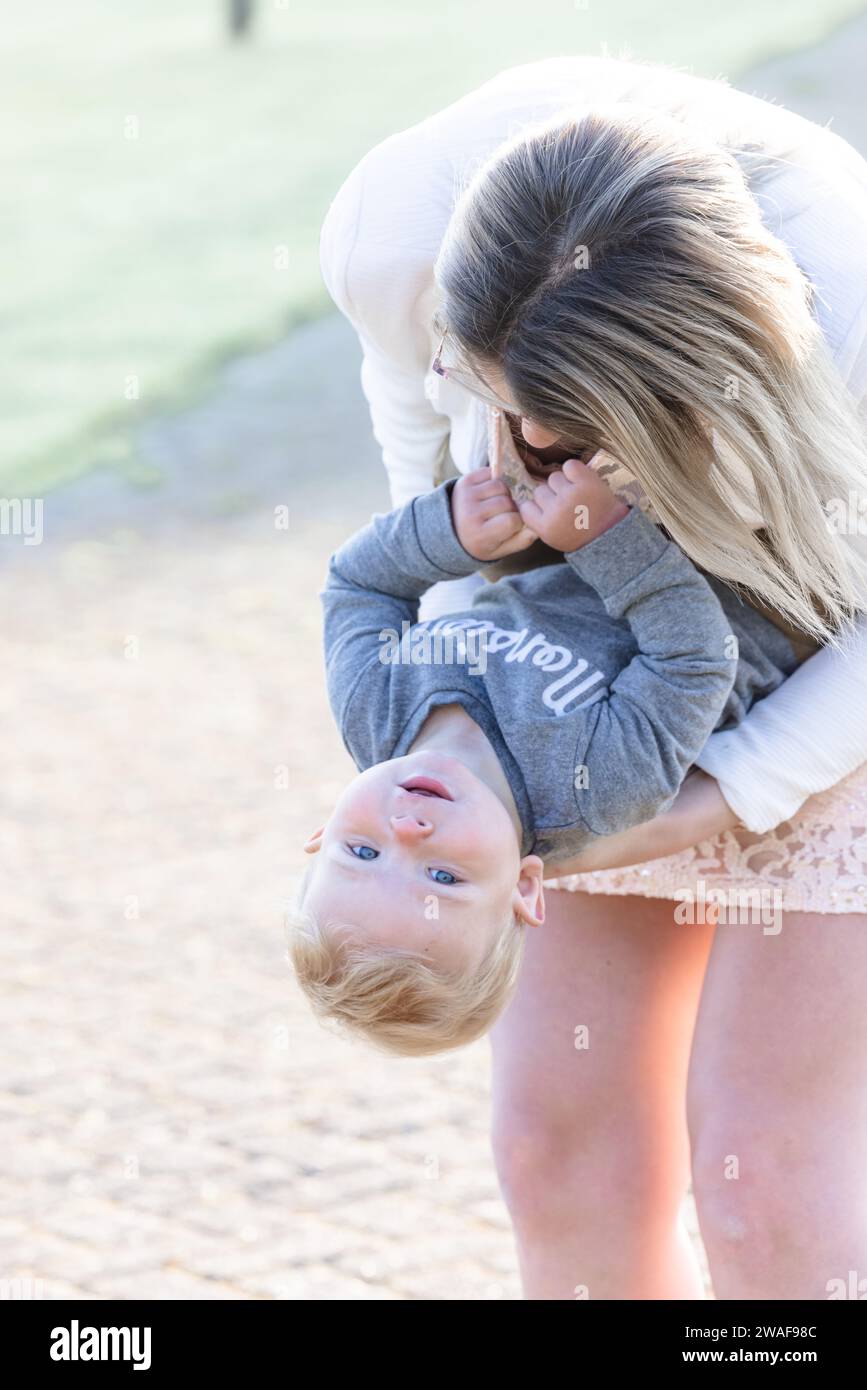 A heartwarming moment is captured here as a young Caucasian woman, with her hair falling forward, lovingly holds a toddler upside down, bringing forth a joyous expression from the child. The soft outdoor lighting gently bathes both, creating a tender atmosphere. The child's bright blue eyes and delighted smile are the focal points of this intimate interaction. Playful Upside-Down Bonding. High quality photo Stock Photo