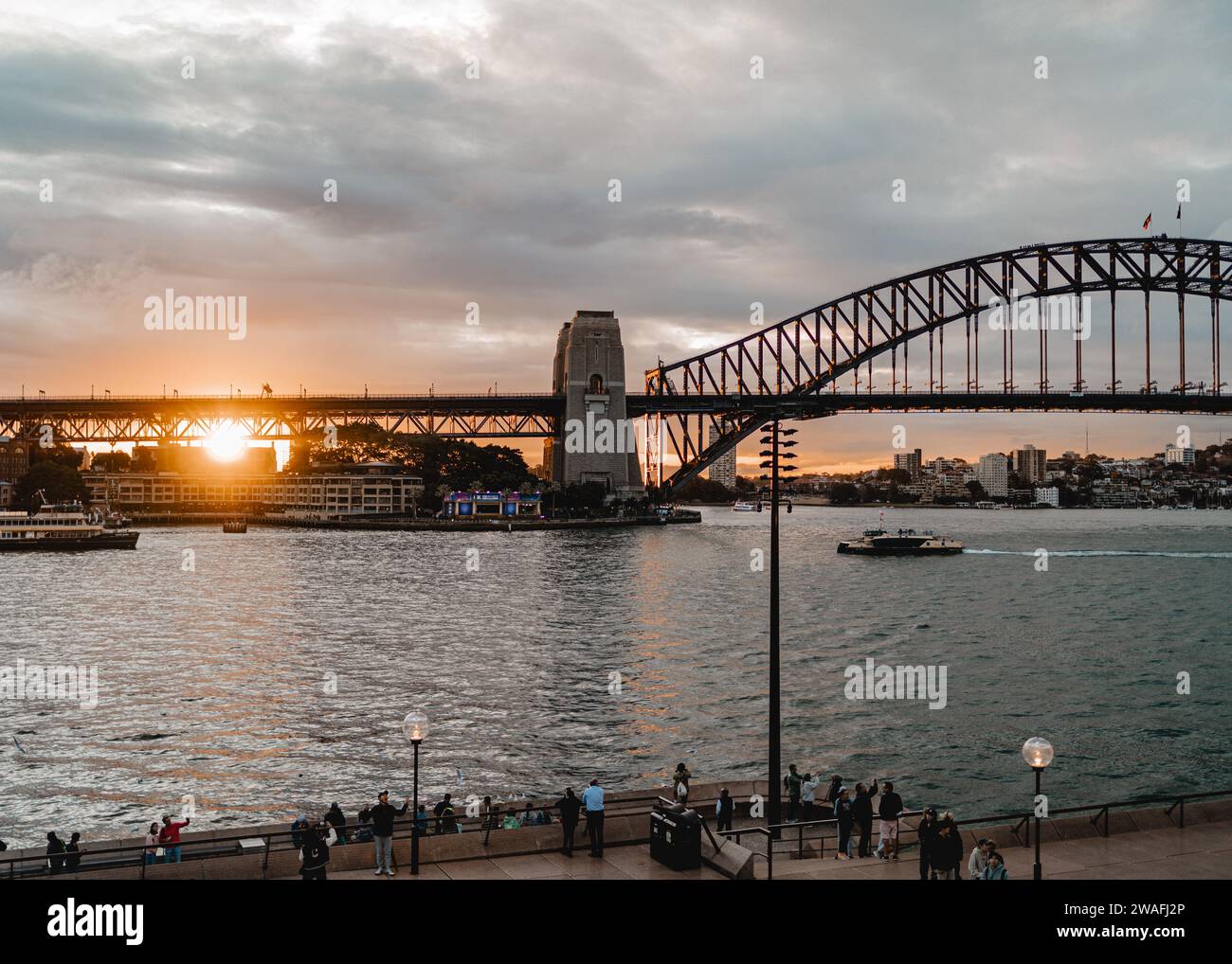 A group of people stand near a bridge that stretches across a body of water,  Sydney Australia Stock Photo