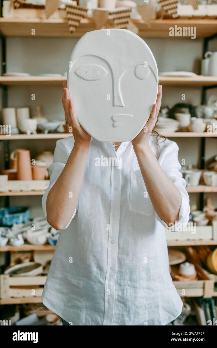 Woman hides her head behind clay product with face relief Stock Photo