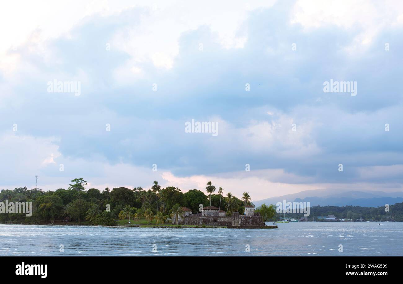 Rio Dulce, Livingston, Guatemala : View of Castillo de San Felipe's ancient walls and towers by the water, encircled by lush palms during a cloudy day Stock Photo