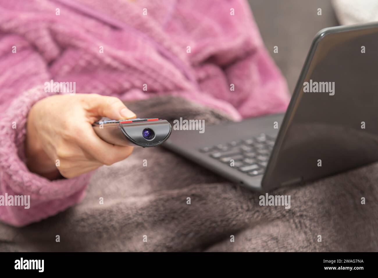 A woman sits on the sofa at home with a TV remote control in her hand and a laptop on her lap. Stock Photo