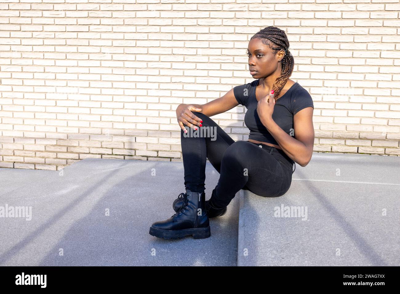 The photograph portrays a young Black woman seated on the concrete ground, leaning against a stark brick wall. Her posture is casual yet composed, with one knee raised and her gaze thoughtfully directed off-camera. She dons an all-black ensemble, from her simple top to her rugged boots, creating a strong visual harmony with her surroundings. The late afternoon sun casts long shadows, adding depth to this scene of urban solitude. Urban Solitude: Reflective Moments in the City. High quality photo Stock Photo