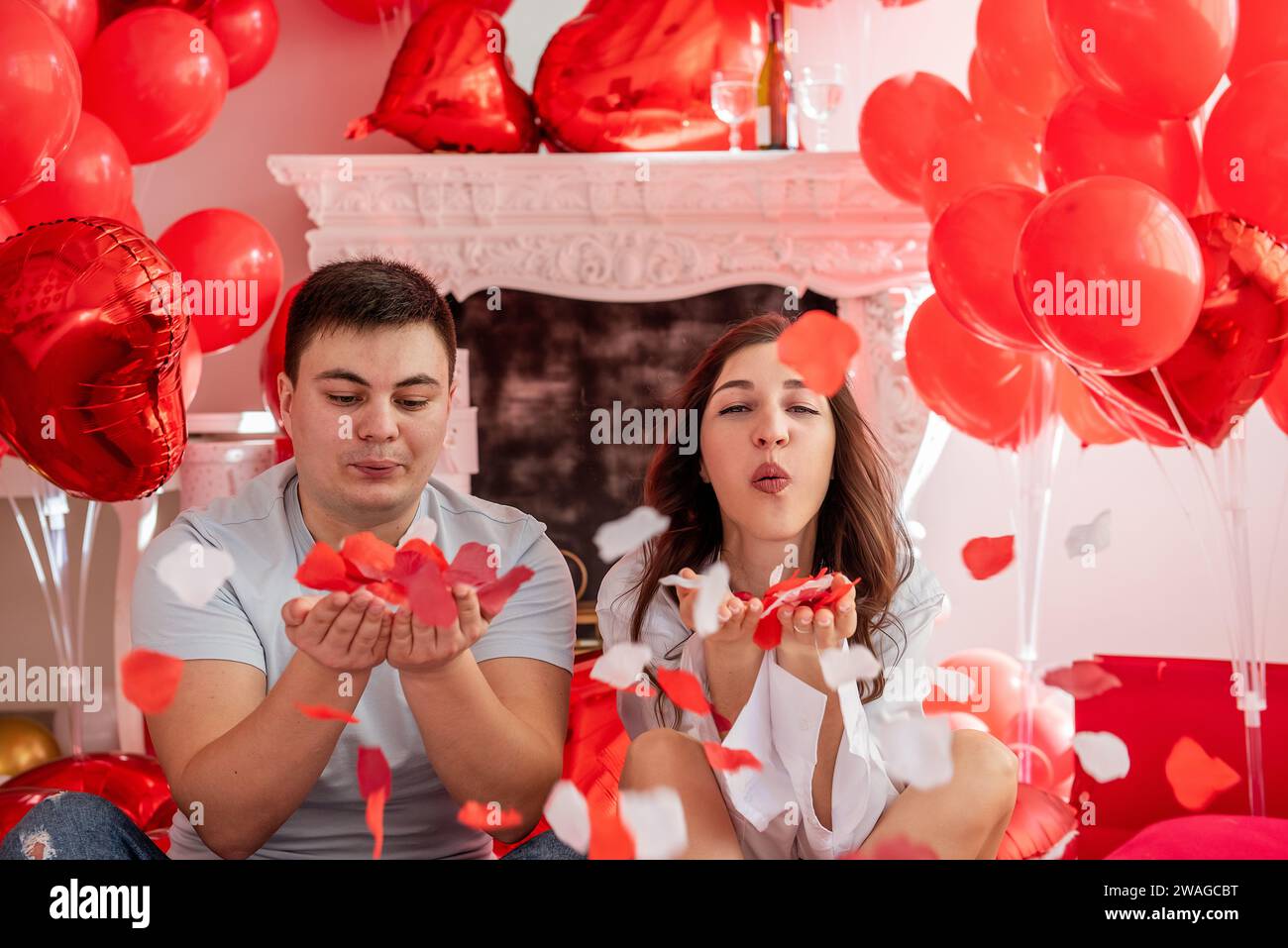 Close up portrait of young man and woman seated close to each other against of white decorative fireplace adorned with red hearts balloons. Couple blo Stock Photo