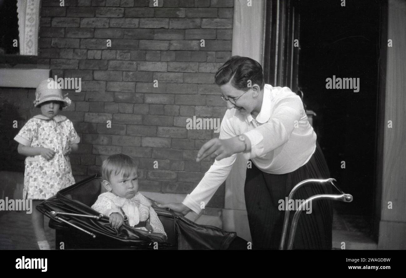 circa 1940s, historical, in the street, outside a house, a happy lady, perhaps an aunt or neighbour, about to lift an infant boy out of a pram of the era, England, UK. In this era and previous eras, it was common for infant children to be left in their prams outside houses and shops, to rest  and sleep and in the believe that fresh air was good for their health. Stock Photo