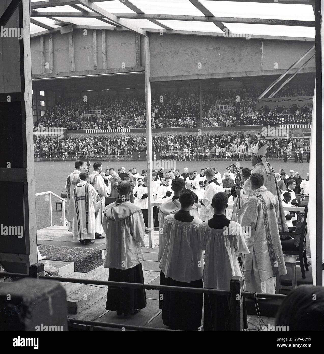 1960s, historical, Catholic pilgrimage, clergy in choir dress and robed priests in pian dress gathered on a temporary wooden stage or platform performing a service inside a stadium, with a large crowd in attendance, England, UK, Stock Photo