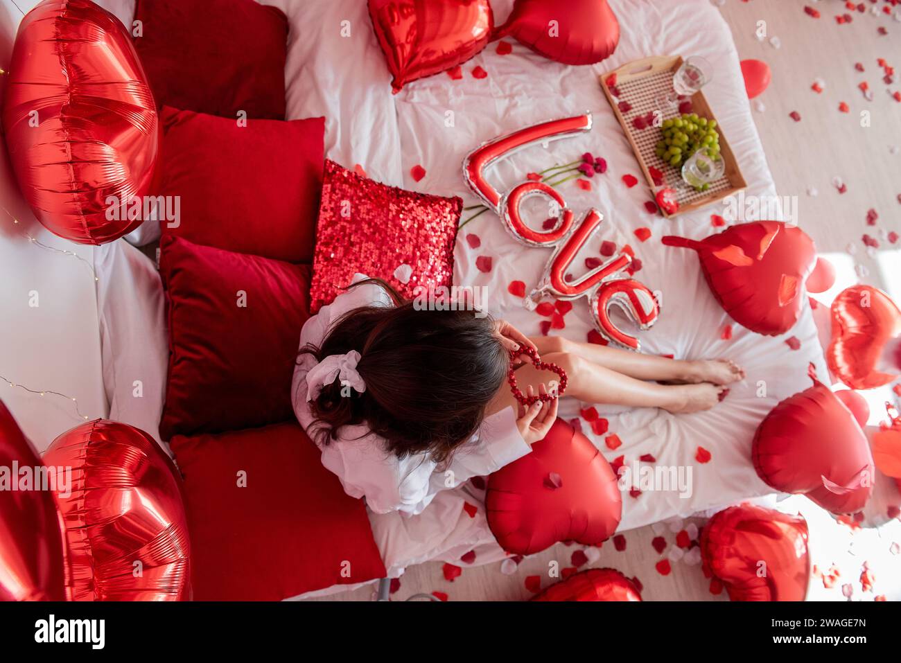 Top view of young woman sitting on bed around red balloons, rose petals. Faceless blogger holds heart-shaped beads. Decoration with inscription Love, Stock Photo
