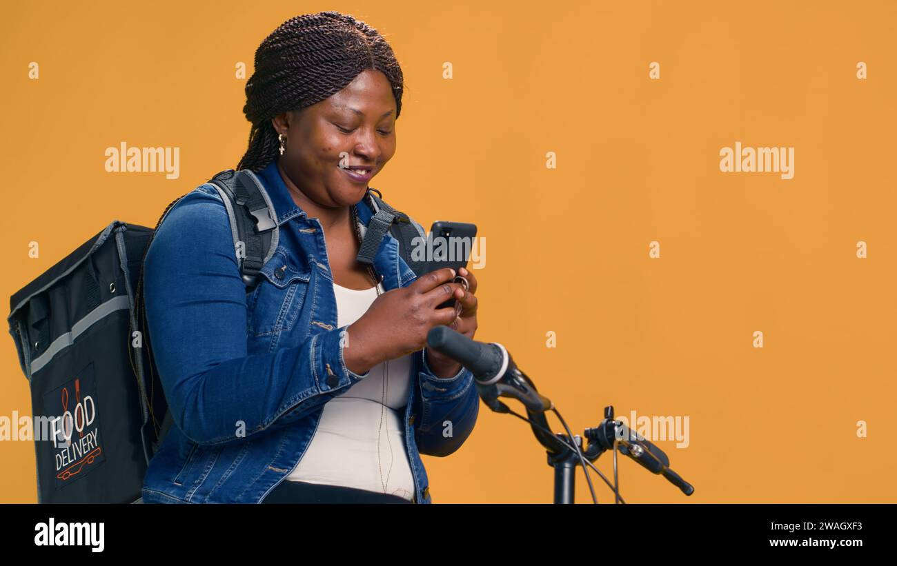 Female bike messenger laughing at message on cell phone while waiting for online order. Enthusiastic african american courier browsing on mobile device while on bicycle. Handheld shot Stock Photo Alam...