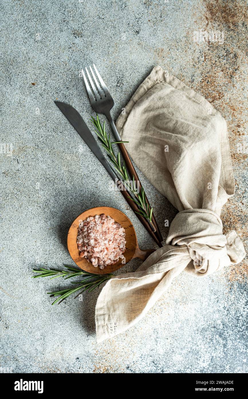 From above wooden spoon with pink Himalayan salt, surrounded by rosemary sprigs and a folded linen napkin alongside a knife and fork, all arranged nea Stock Photo