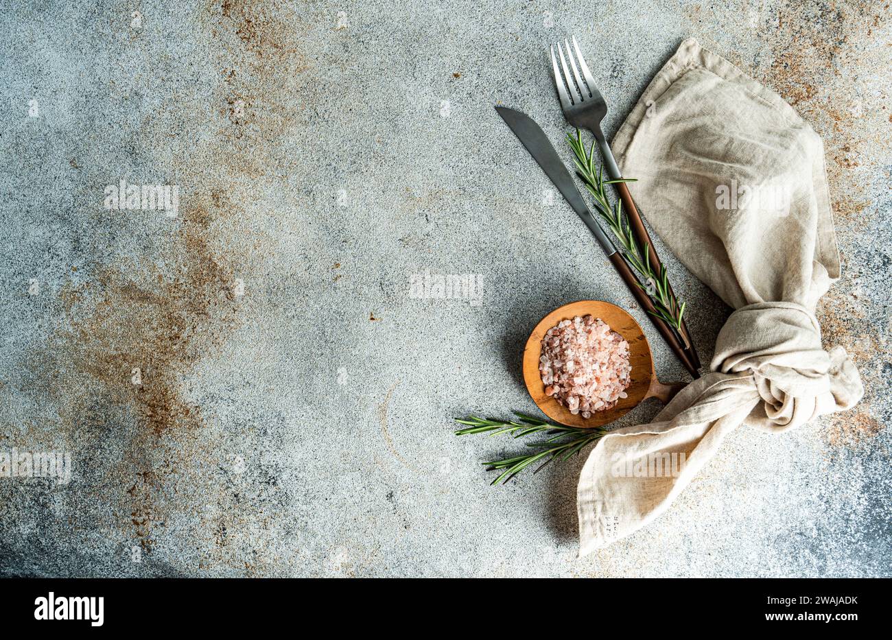 From above wooden spoon with pink Himalayan salt, surrounded by rosemary sprigs and a folded linen napkin alongside a knife and fork, all arranged nea Stock Photo