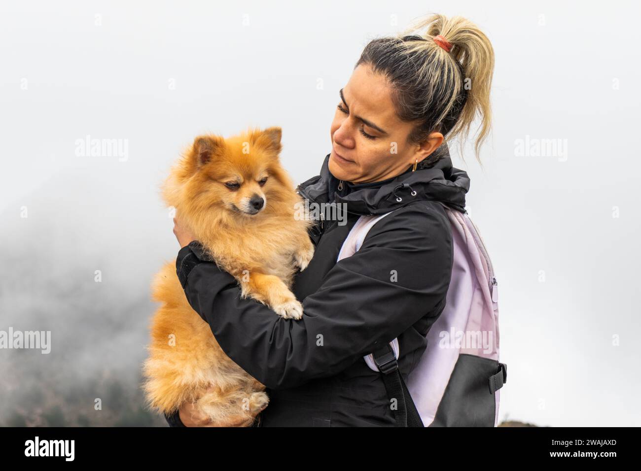 A woman lovingly holds her Pomeranian dog, both enjoying the outdoors with a foggy mountain backdrop in Cumbres del Ajusco National Park Stock Photo