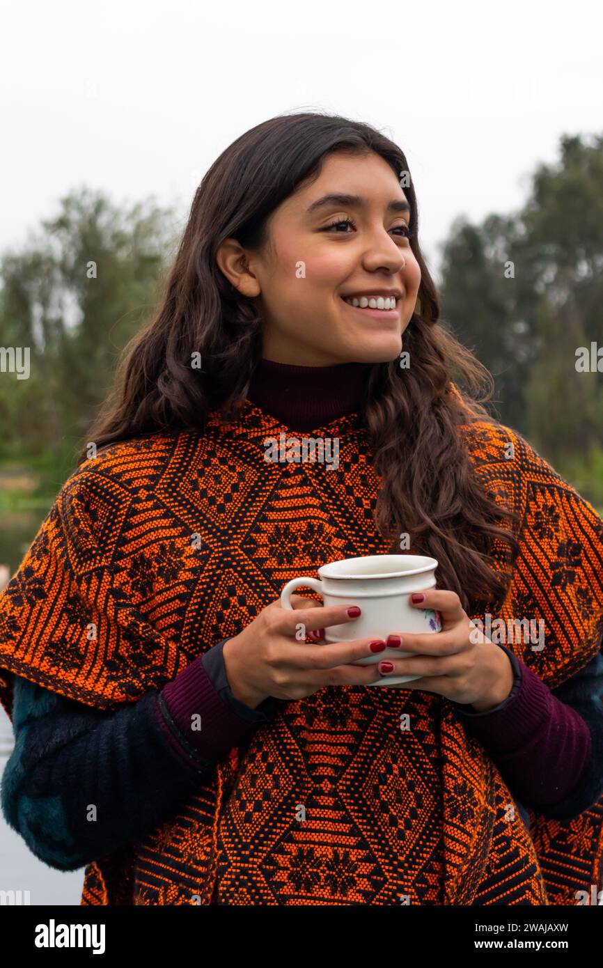 Portrait joyful woman wrapped in a traditional patterned shawl holds a mug smiling outdoors with a soft focus background Stock Photo