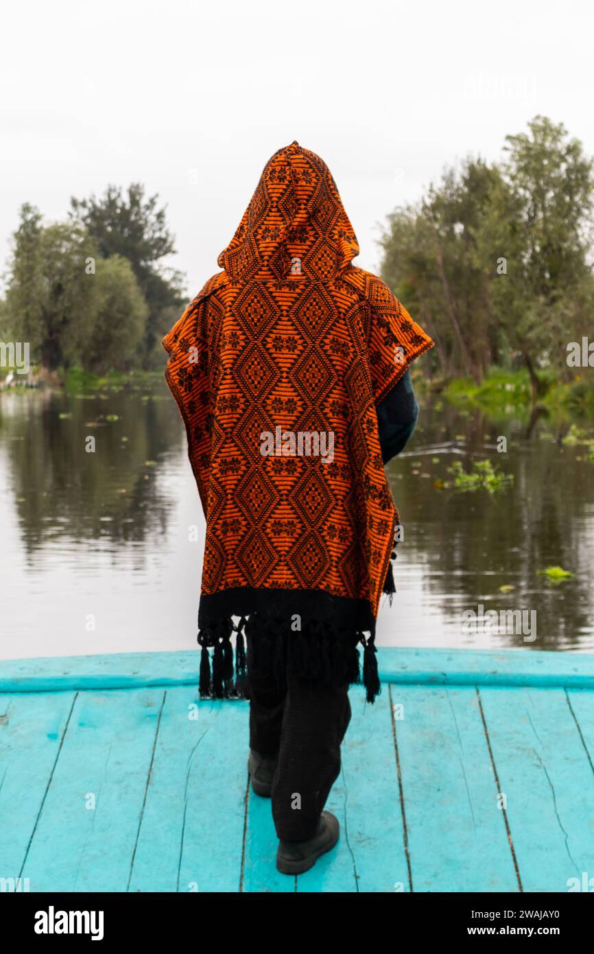 Back view of person draped in an ornate shawl stands facing the lush scenery of Xochimilcos waterways Stock Photo