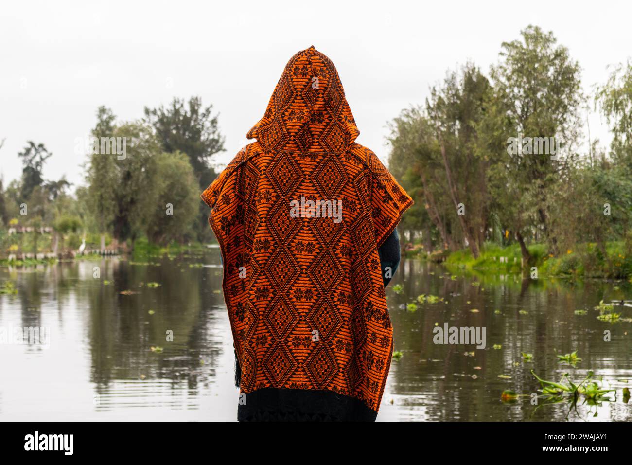 Back view of person draped in an ornate shawl stands facing the lush scenery of Xochimilcos waterways Stock Photo