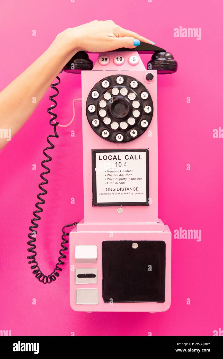 Crop anonymous woman's hand delicately touching a vintage pink rotary wall telephone, set against a contrasting pink background Stock Photo