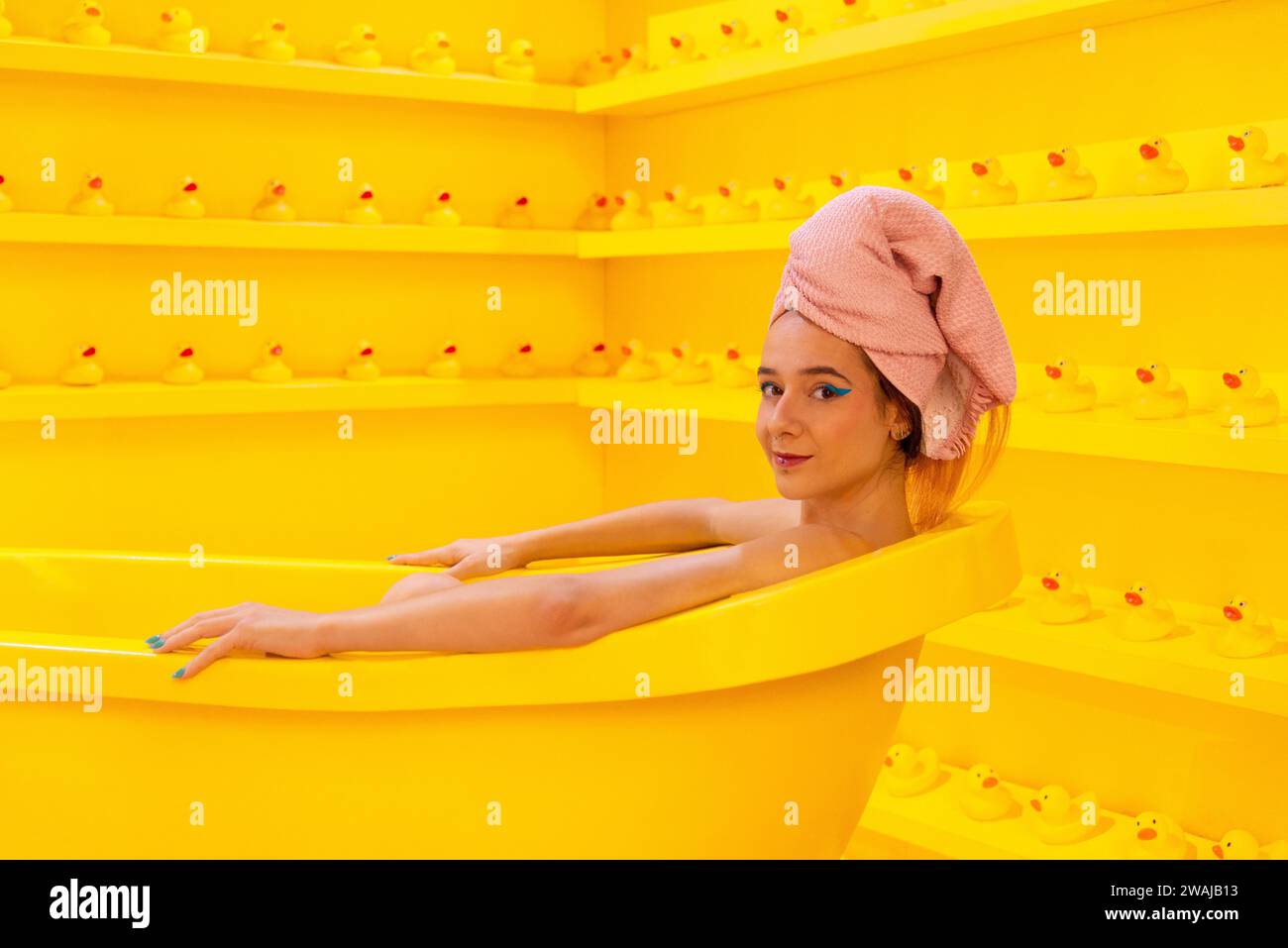 A woman with a towel wrapped head looking at camera relaxing in a bright yellow bathtub surrounded by rubber ducks in a monochromatic setting Stock Photo