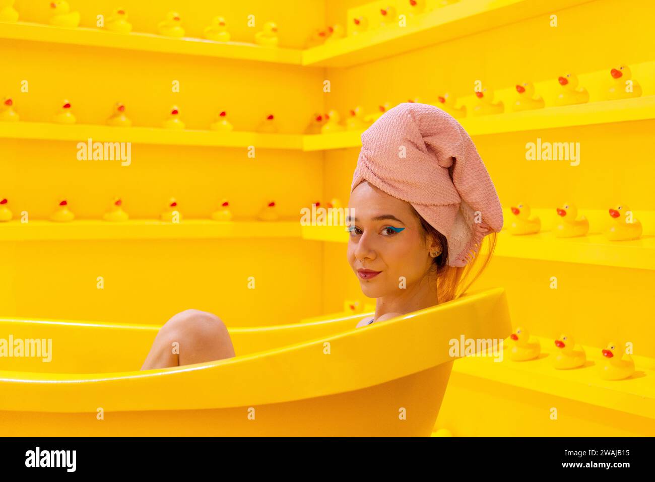 A woman with a towel wrapped head relaxing in a bright yellow bathtub surrounded by rubber ducks in a monochromatic setting Stock Photo