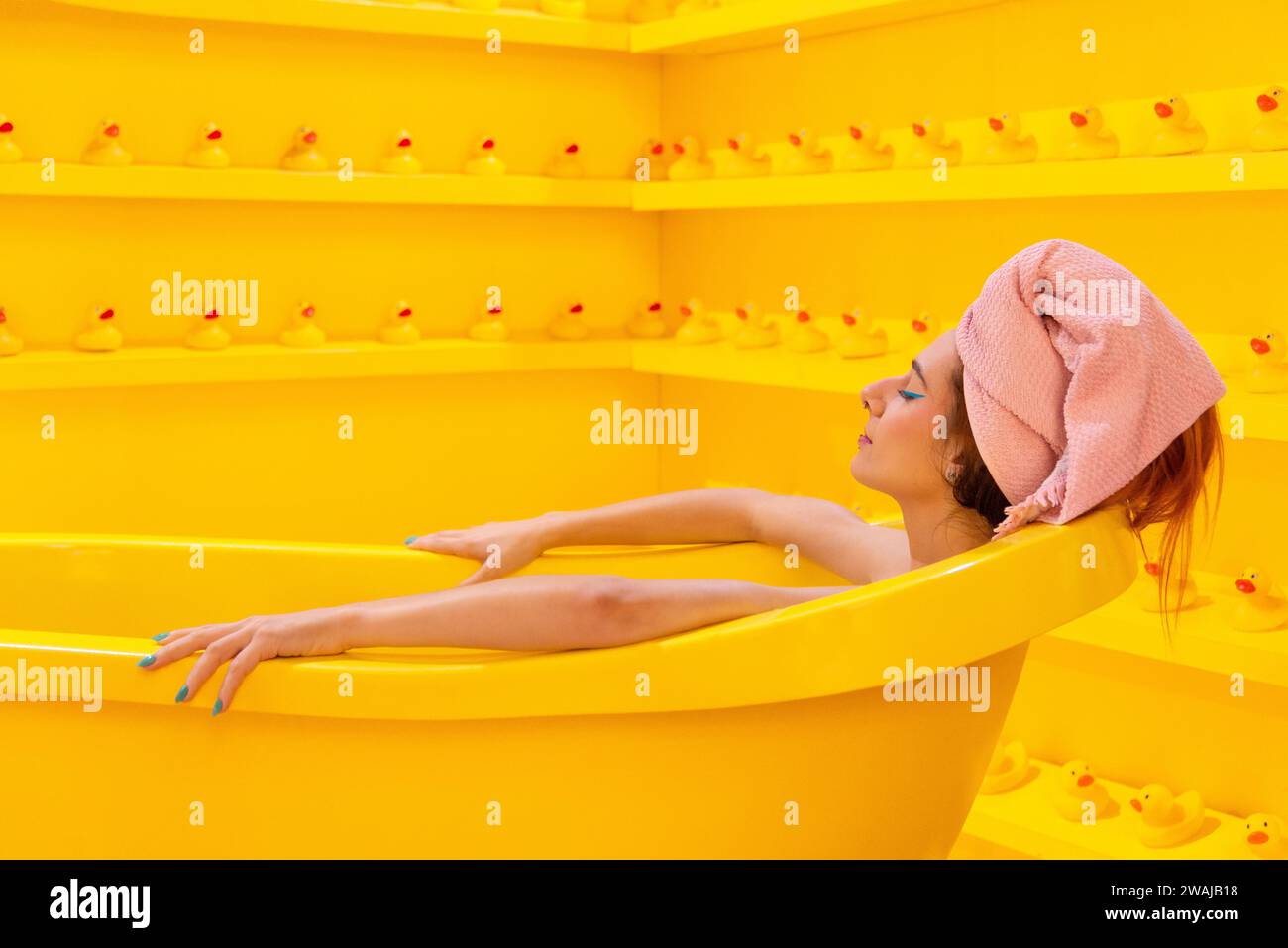 Woman relaxing in a yellow bathtub surrounded by rubber ducks in a monochromatic yellow room Stock Photo