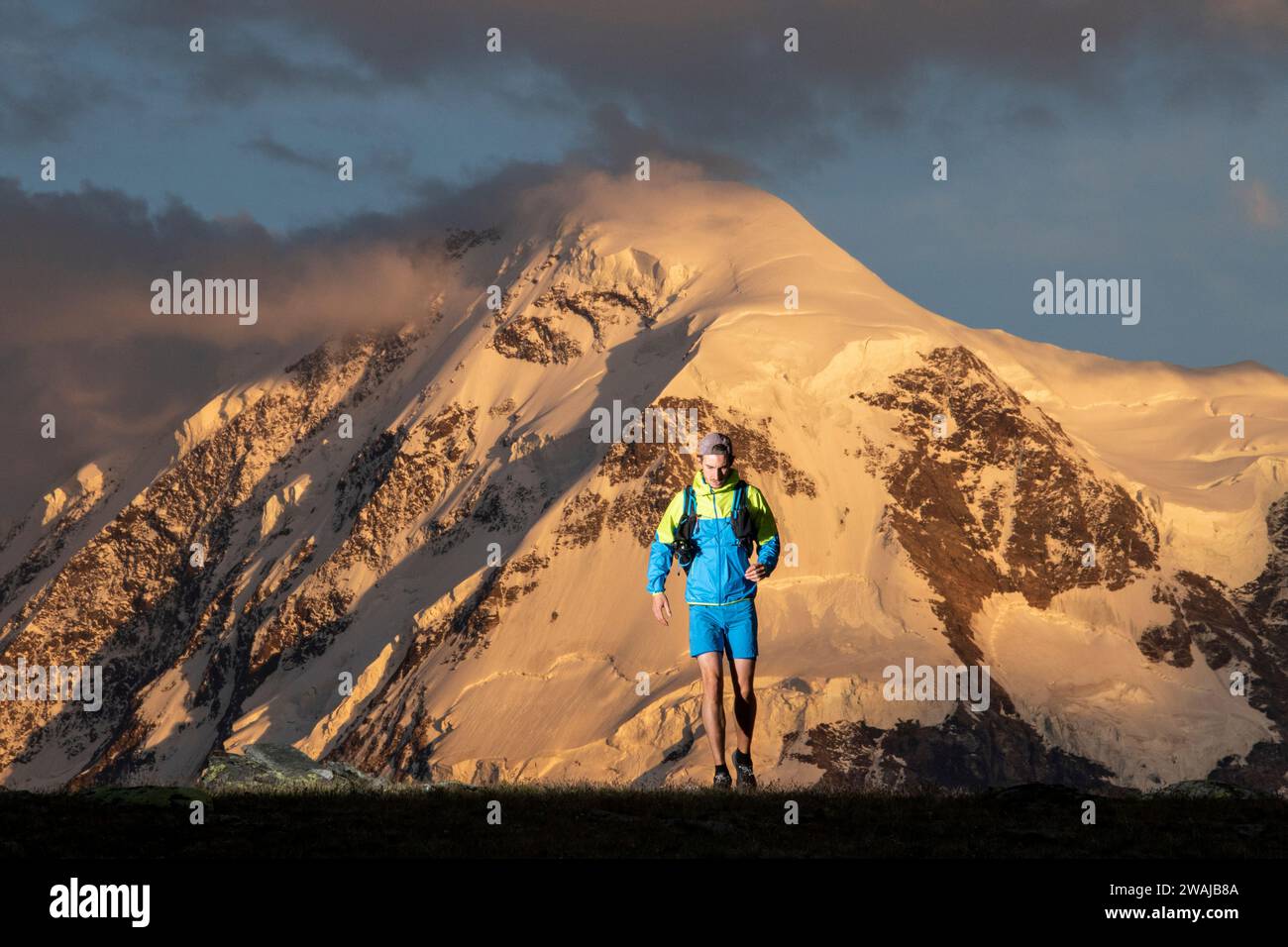 A hiker with backpack treks against a backdrop of majestic snow-covered mountains bathed in sunset light Stock Photo