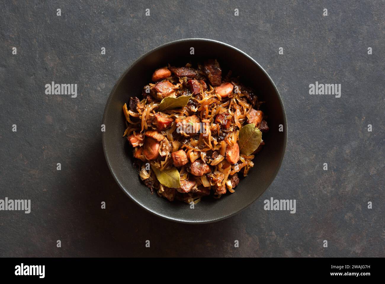 Polish Bigos in bowl over dark stone background. Stewed cabbage with sauerkraut, mushrooms, smoked meats and spices. Top view, flat lay Stock Photo