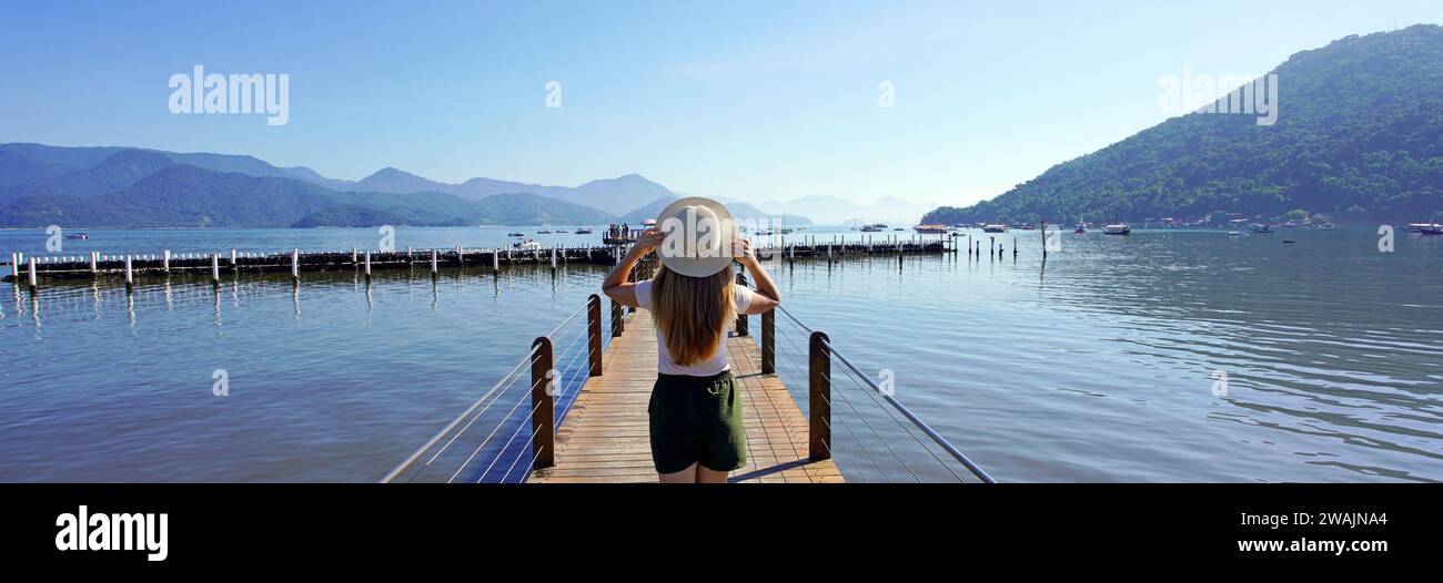 Holidays in Brazil. Panoramic banner view of young traveler woman walking on pier enjoying landscape in Ubatuba, Brazil. Stock Photo