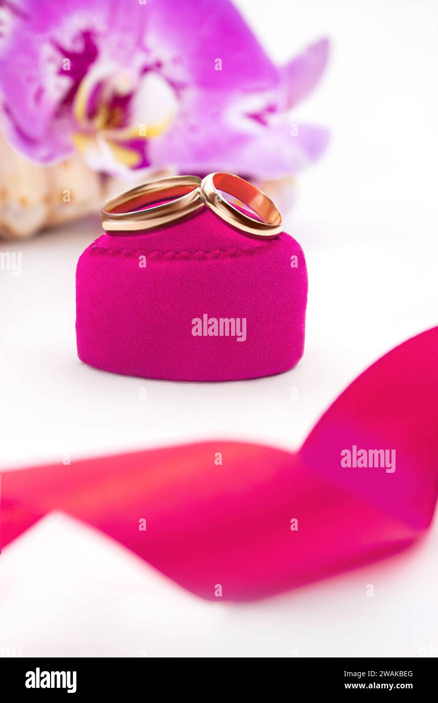 Gold wedding rings on pink velvet heart on gift box with orchids on white background. Marriage, proposal, Valentine's Day. Vertical. Copy space Stock Photo