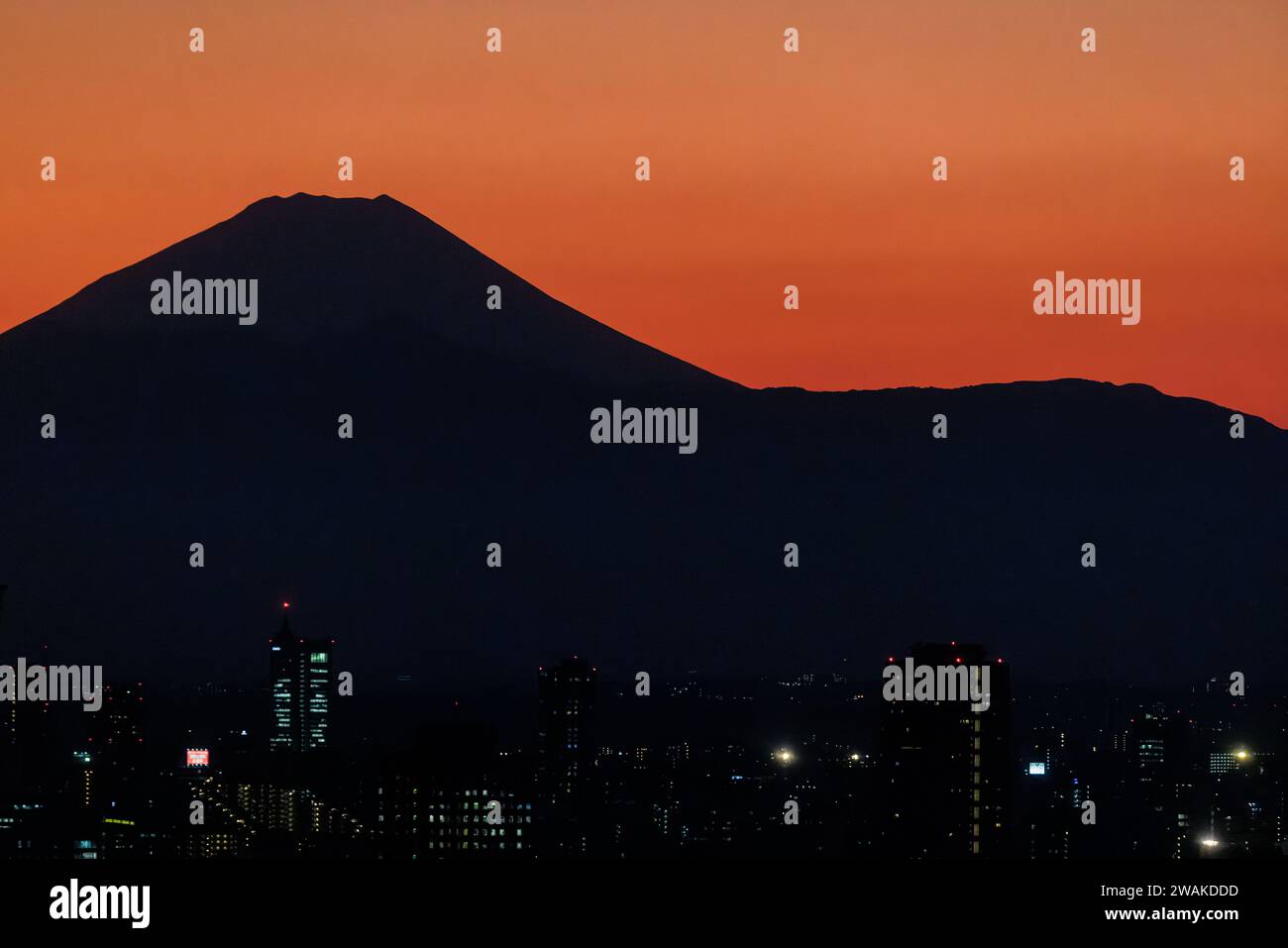 black silhouette of the volcanic cone of mount fuji set against a bright orange sunset with tokyo city lights in the foreground Stock Photo
