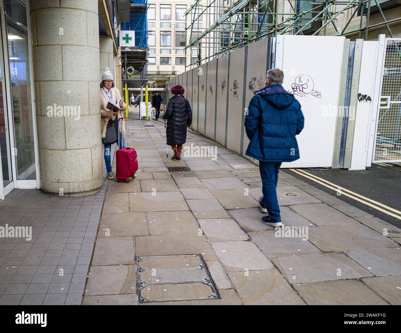 a woman stands reading, with her luggage by her feet, while other people pass by Stock Photo