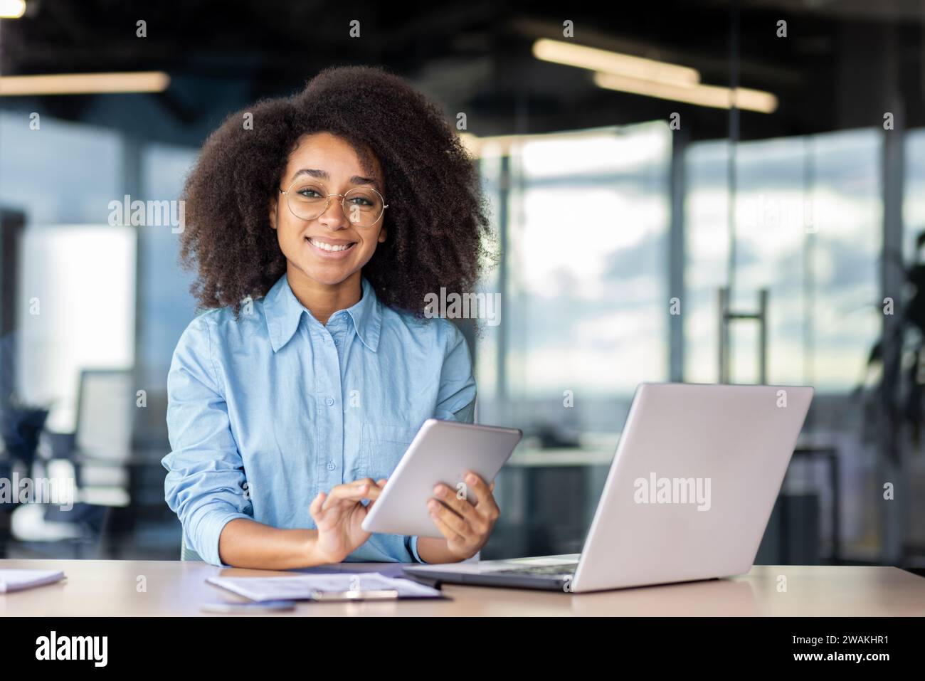 Portrait of a smiling young African American woman sitting at a desk in the office, holding a tablet and smiling at the camera. Stock Photo