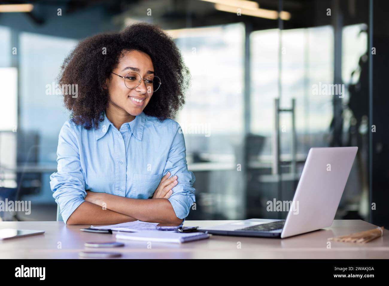 A smiling young African-American woman is sitting in the office at the desk and looking at the laptop screen. Works, studies, talks via video call. Stock Photo