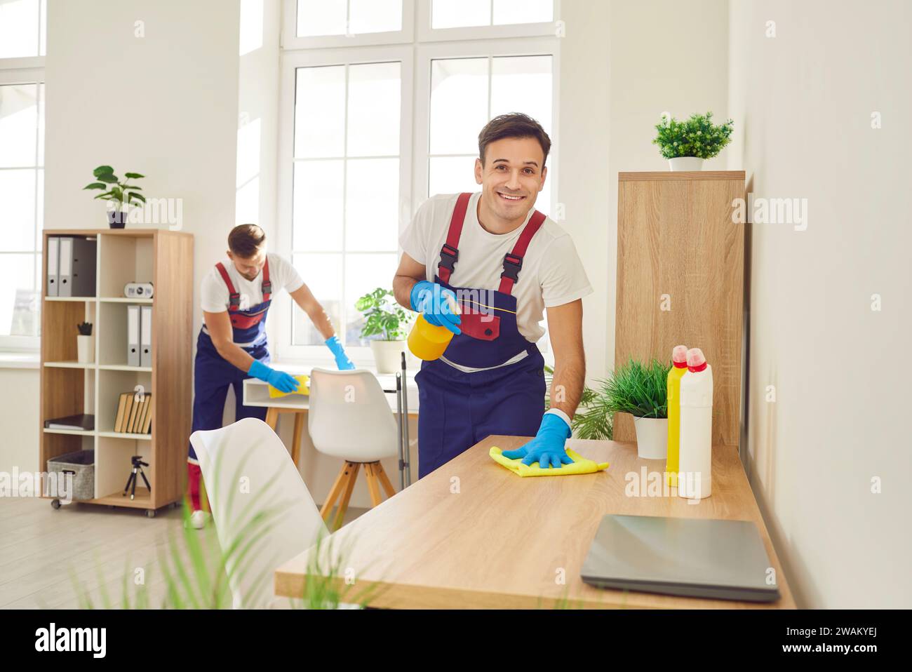 Two happy professional janitors in uniforms cleaning office tables with sanitizer Stock Photo