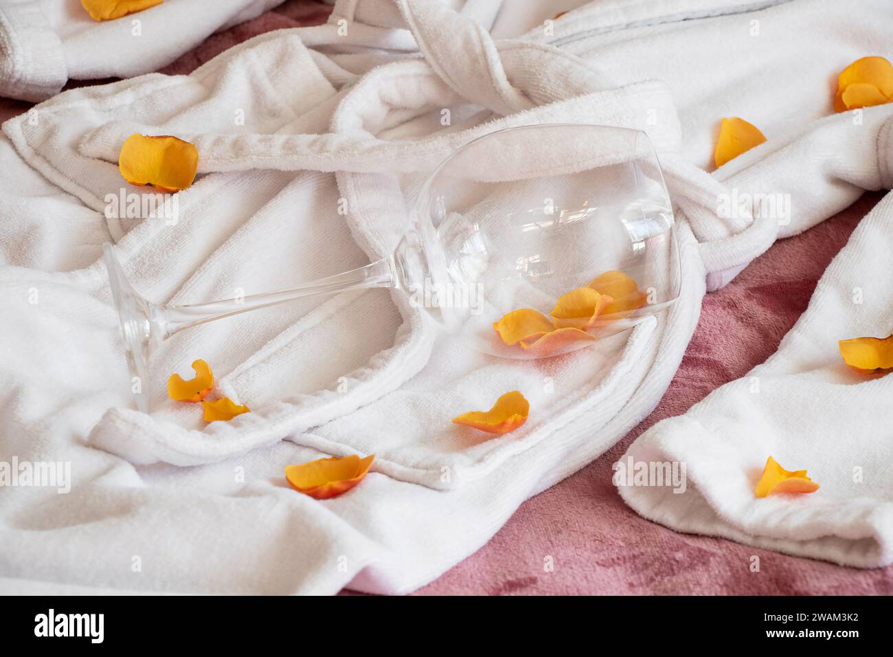 A woman's white terry robe lies on the bed, rose petals are scattered and an empty glass of wine, romantic relationship Stock Photo