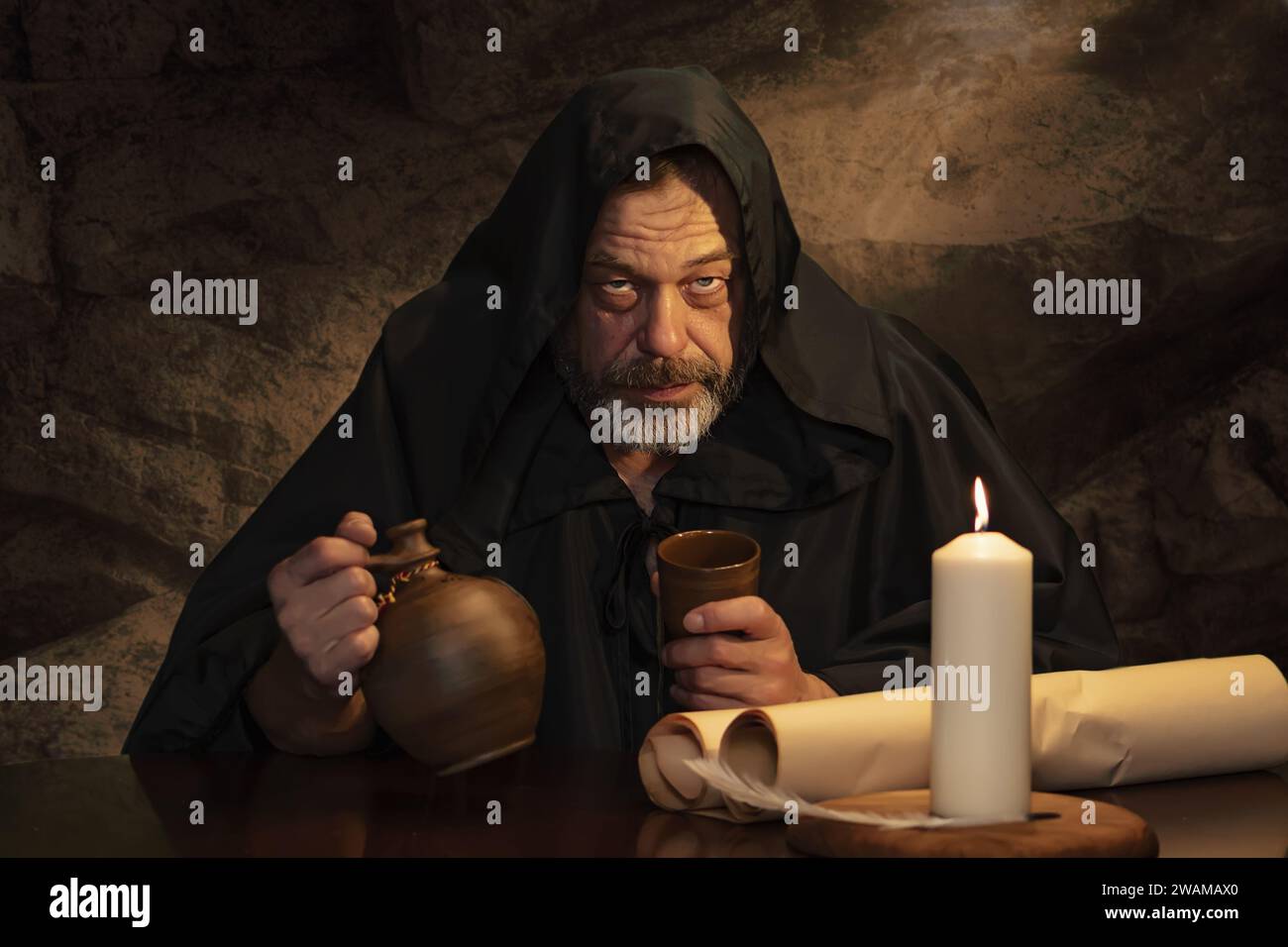 Emotional portrait of a monk in cassock, clay goblet in hand, sitting at a table, a jug of wine and drinking in a medieval inn. Concept: drunkenness a Stock Photo
