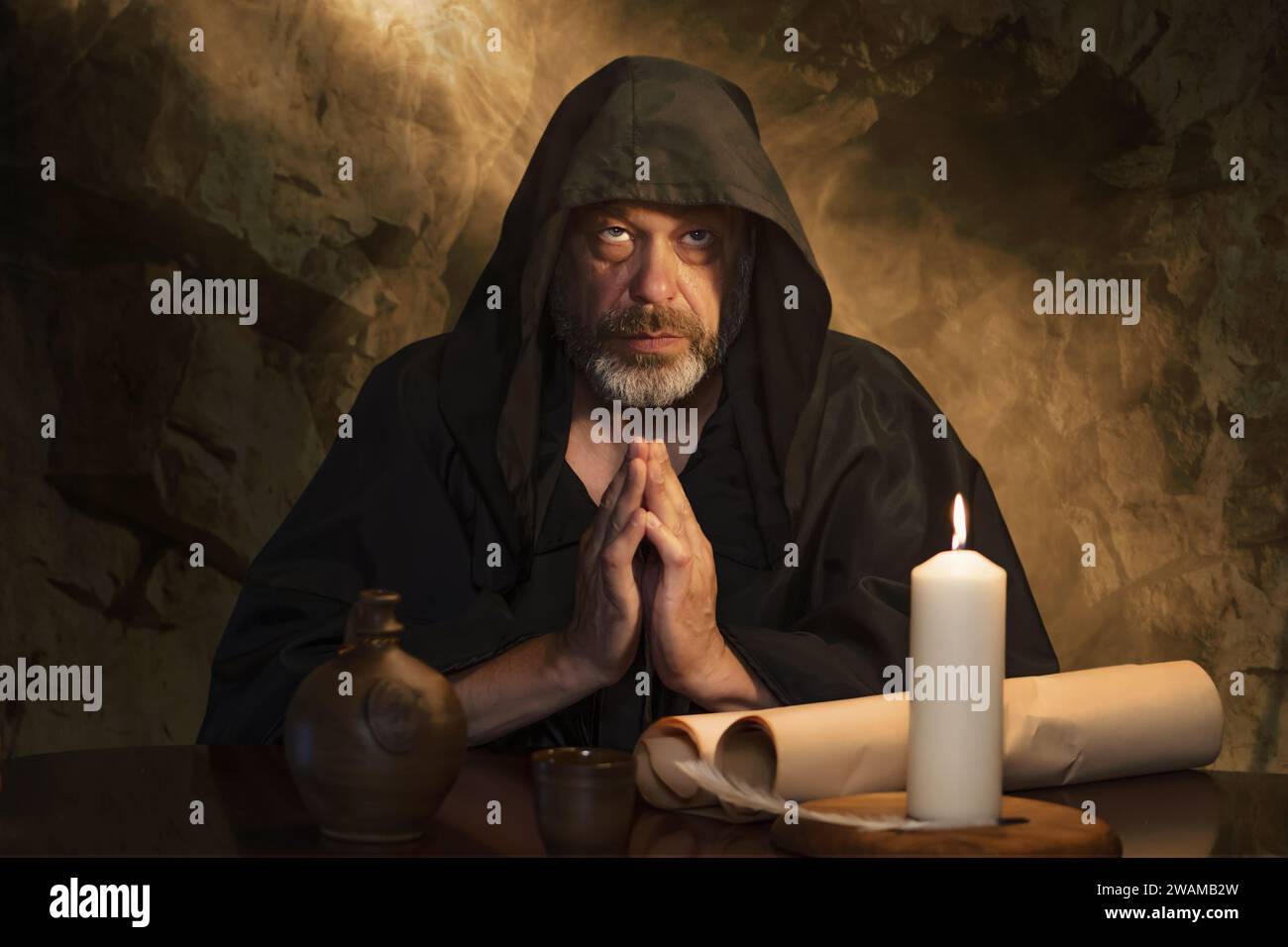 portrait of an elderly monk in a black robe, hands folded in prayer, a dark cell, a candle burning on a table. Stock Photo