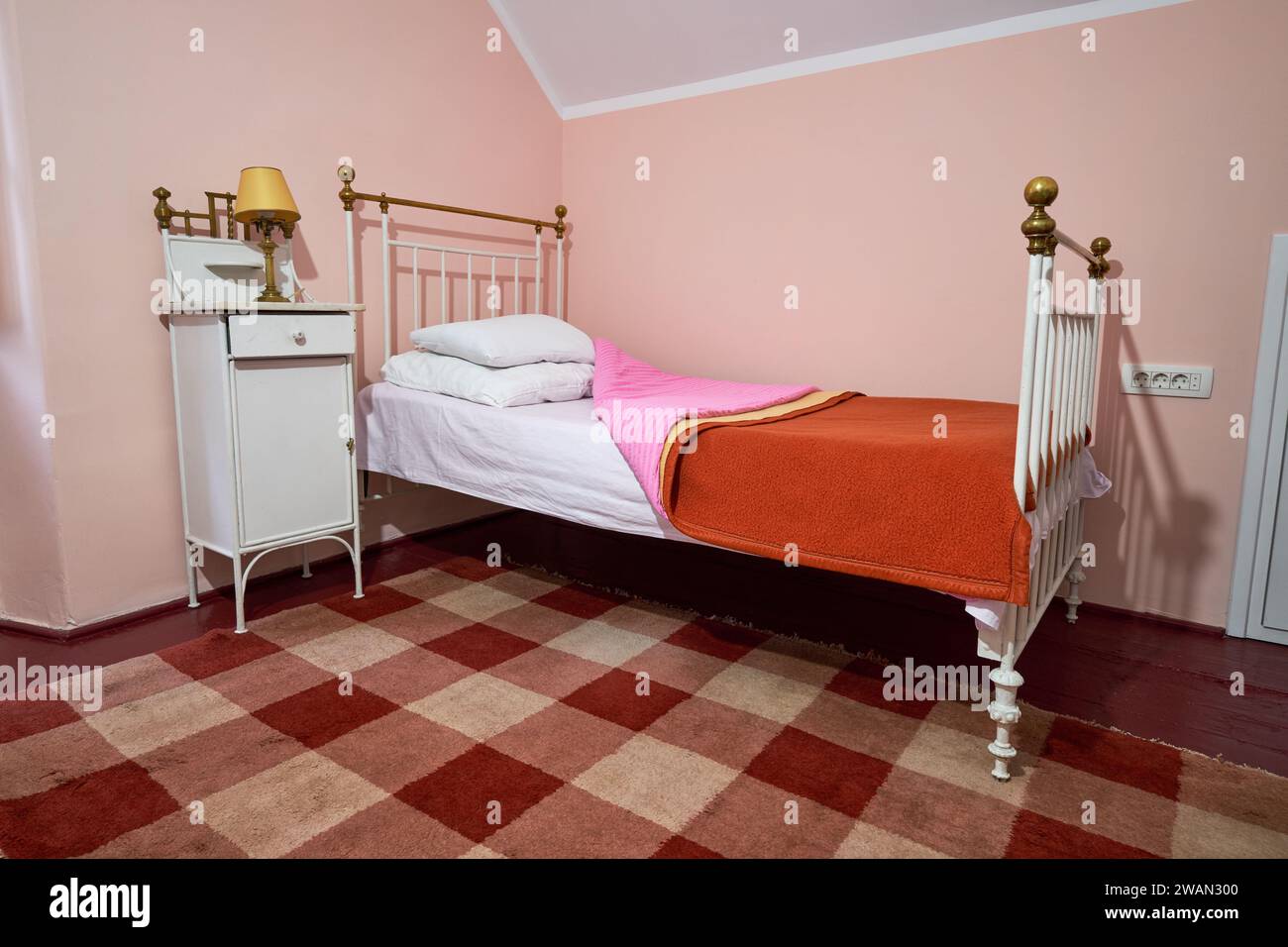 Old cabin type bedroom with white iron bed, rustic pink and orange bed covers and checkered rug. Stock Photo
