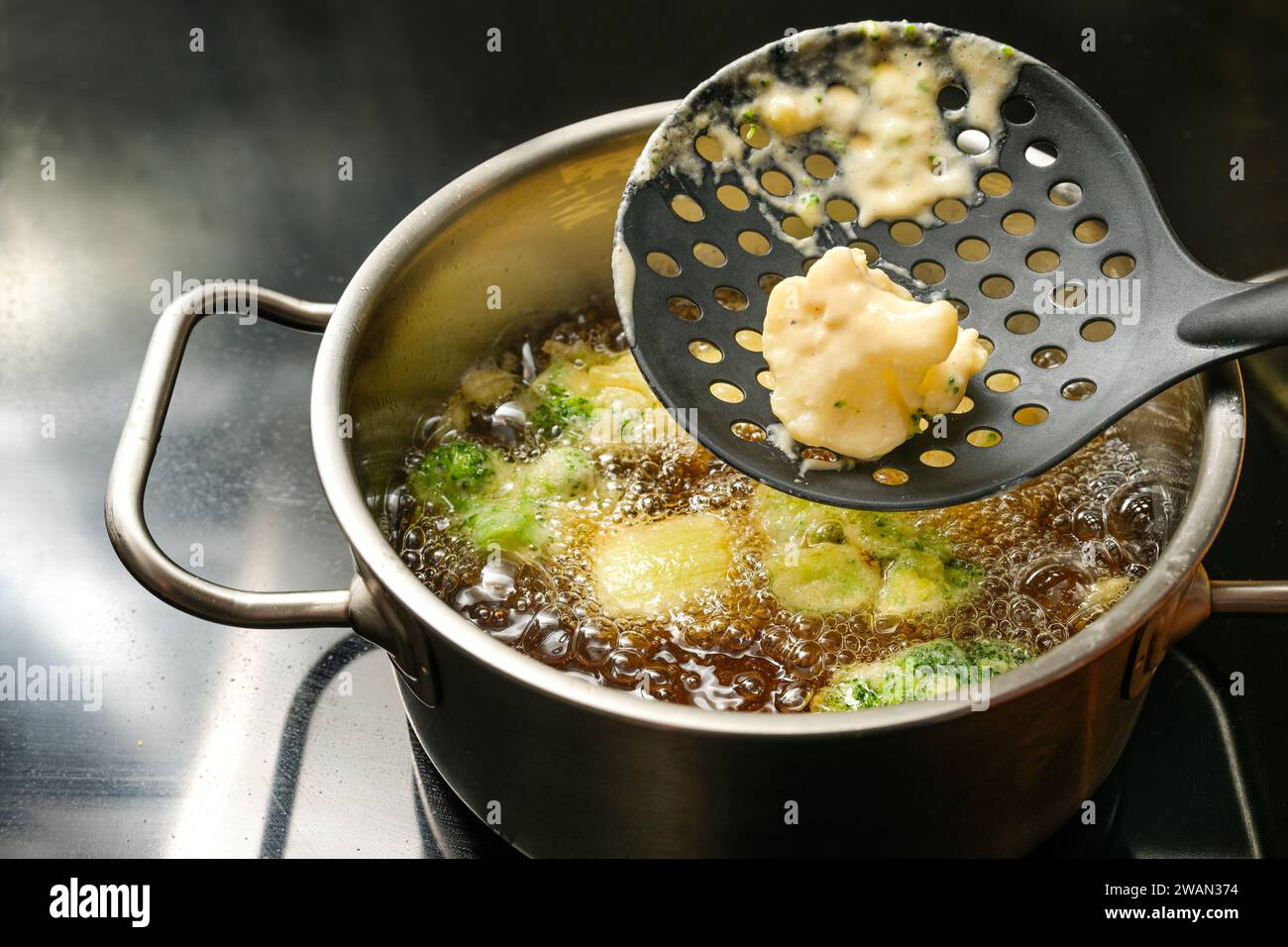 Deep frying cauliflower and broccoli wrapped with beer batter in a pot with hot boiling oil on a black stove top, cooking vegan fast food with vegetab Stock Photo
